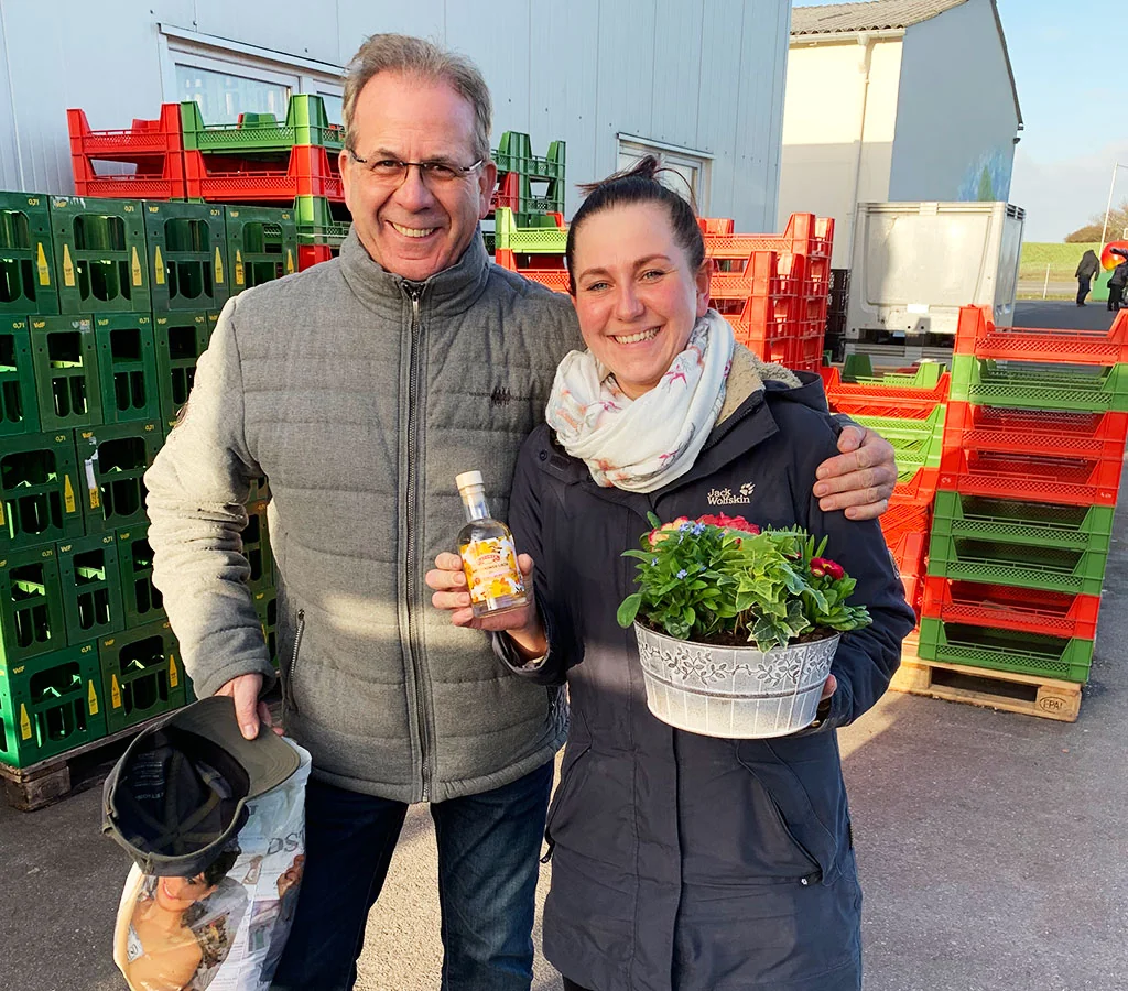 Zwei Kolleginnen und Kollegen mit Blumentopf und kleiner Flasche vor Getränkekisten