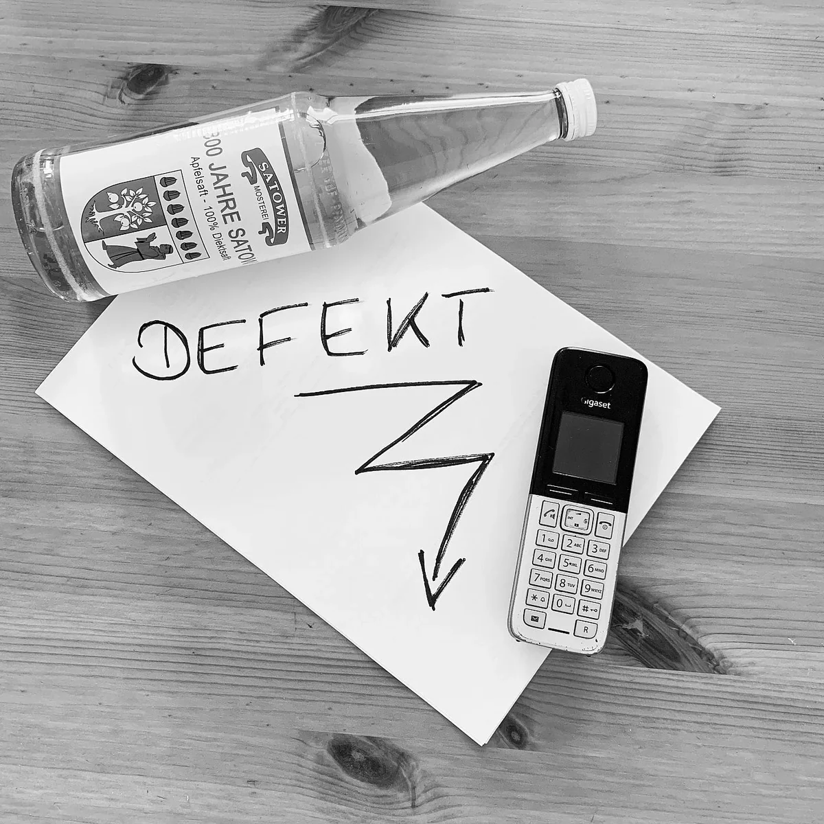 Schwarzweißfoto mit „Defekt“-Zettel neben Telefon und Saftflasche