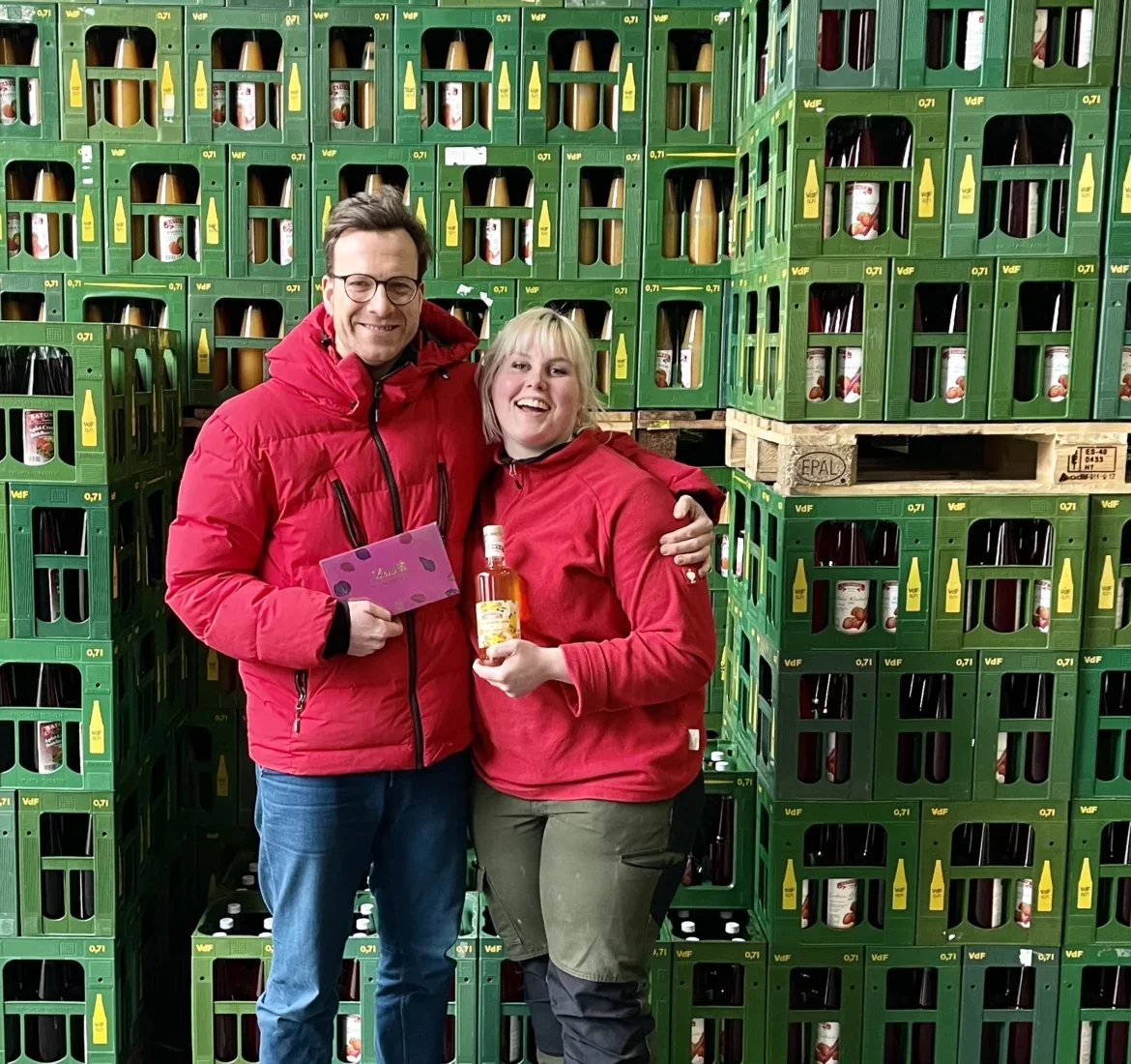 Zwei Mitarbeitende mit Geschenk und Flasche vor gestapelten Saftkisten