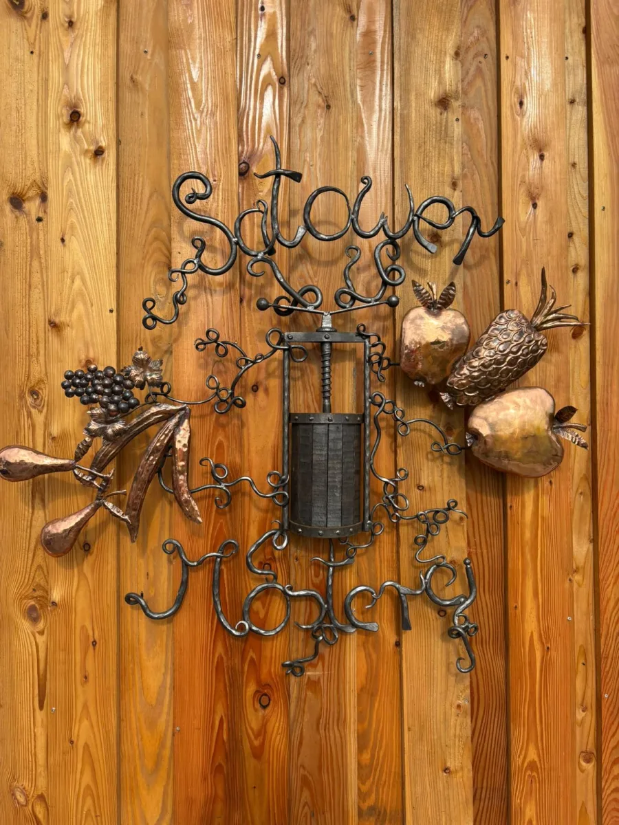 Geschmiedetes Schild „Satower Mosterei“ auf Holzwand: eiserner Schriftzug um eine kleine Spindelpresse, dazu Früchte aus Kupfer